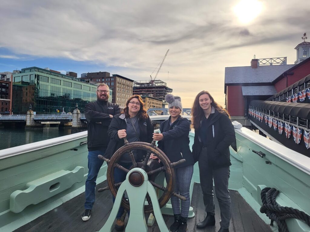Four people posing on a boat deck with a steering wheel under a cloudy sky.