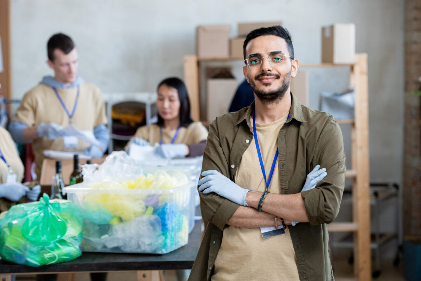 Gloved young man crossing arms by chest against volunteers sorting garbage