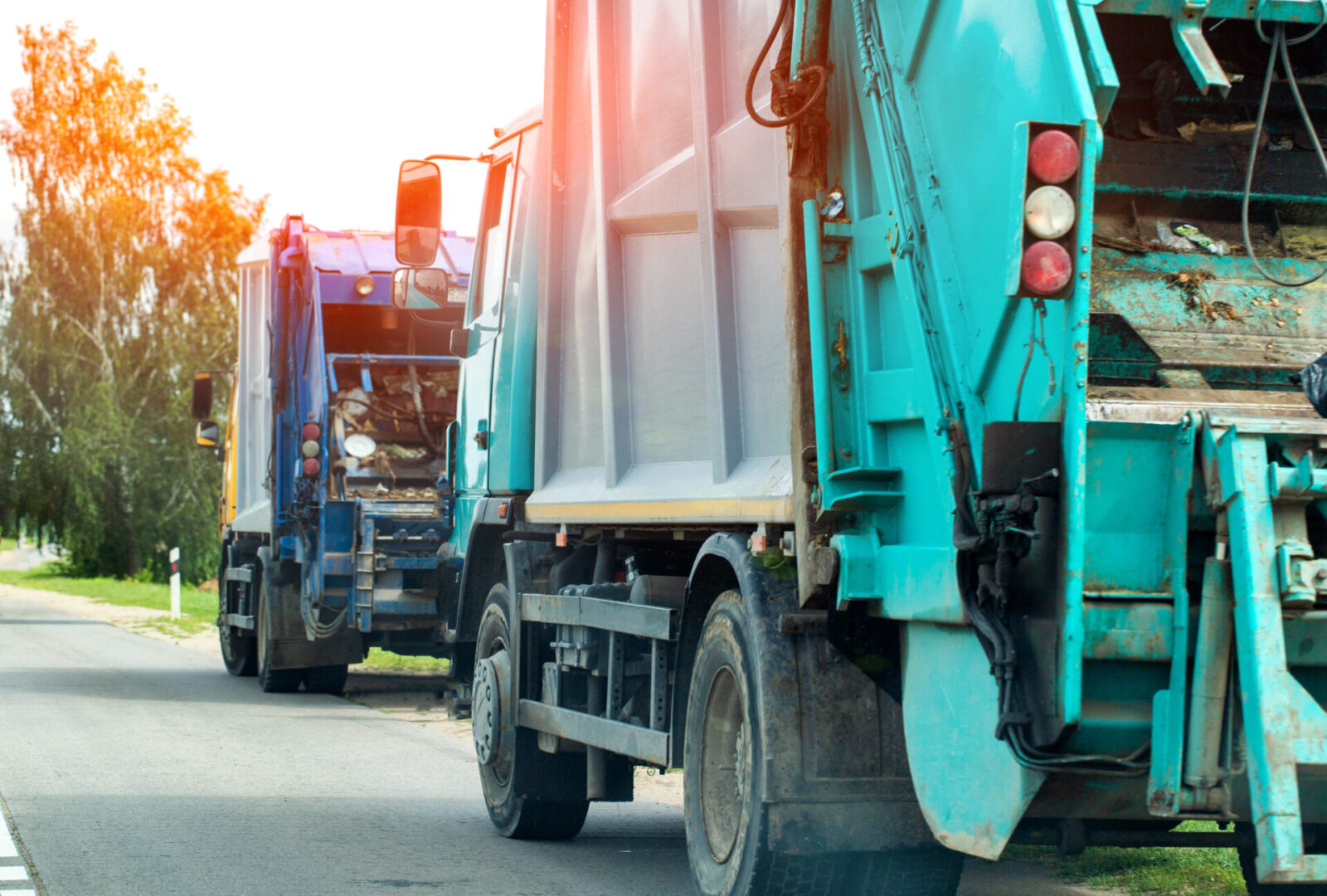 Side and rear view of two turquoise and blue garbage trucks on asphalt road, green trees, daytime garbage collection.